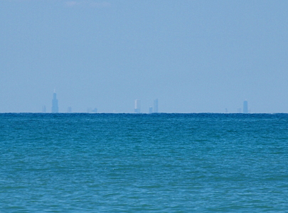 [A lake view with four very tall buildings and a number of smaller ones visible behind the haze at the water's surface.]
